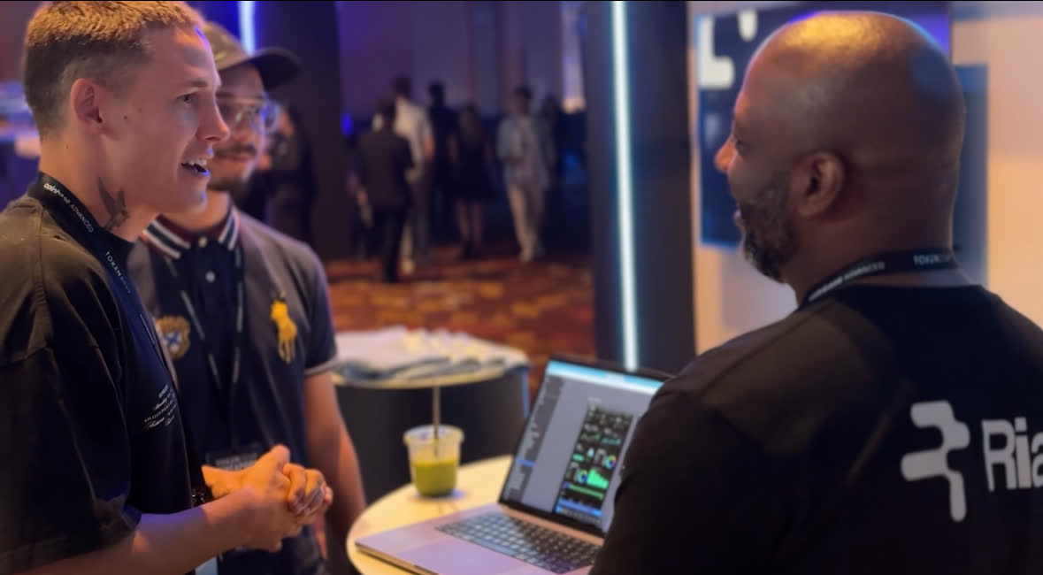 Three people talking across a table on a conference floor, a laptop open between them.